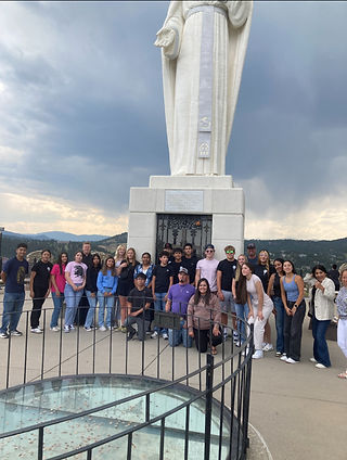 Group of people posing in front of a large statue with a mountainous backdrop and cloudy sky. The mood is cheerful.