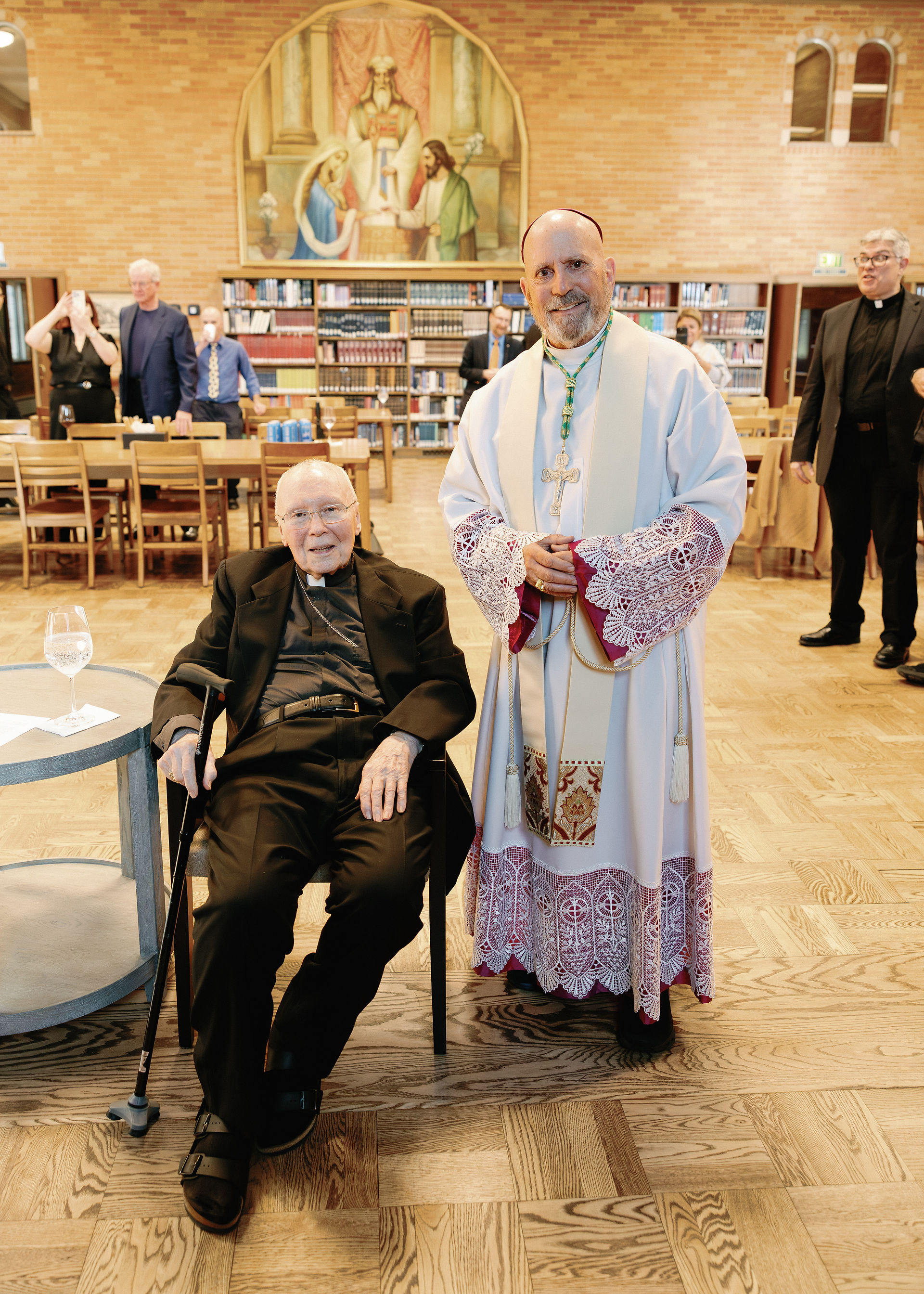 Two clergymen, one seated with a cane, another in white robes, pose in a library. A painting and onlookers in the background. Warm mood.
