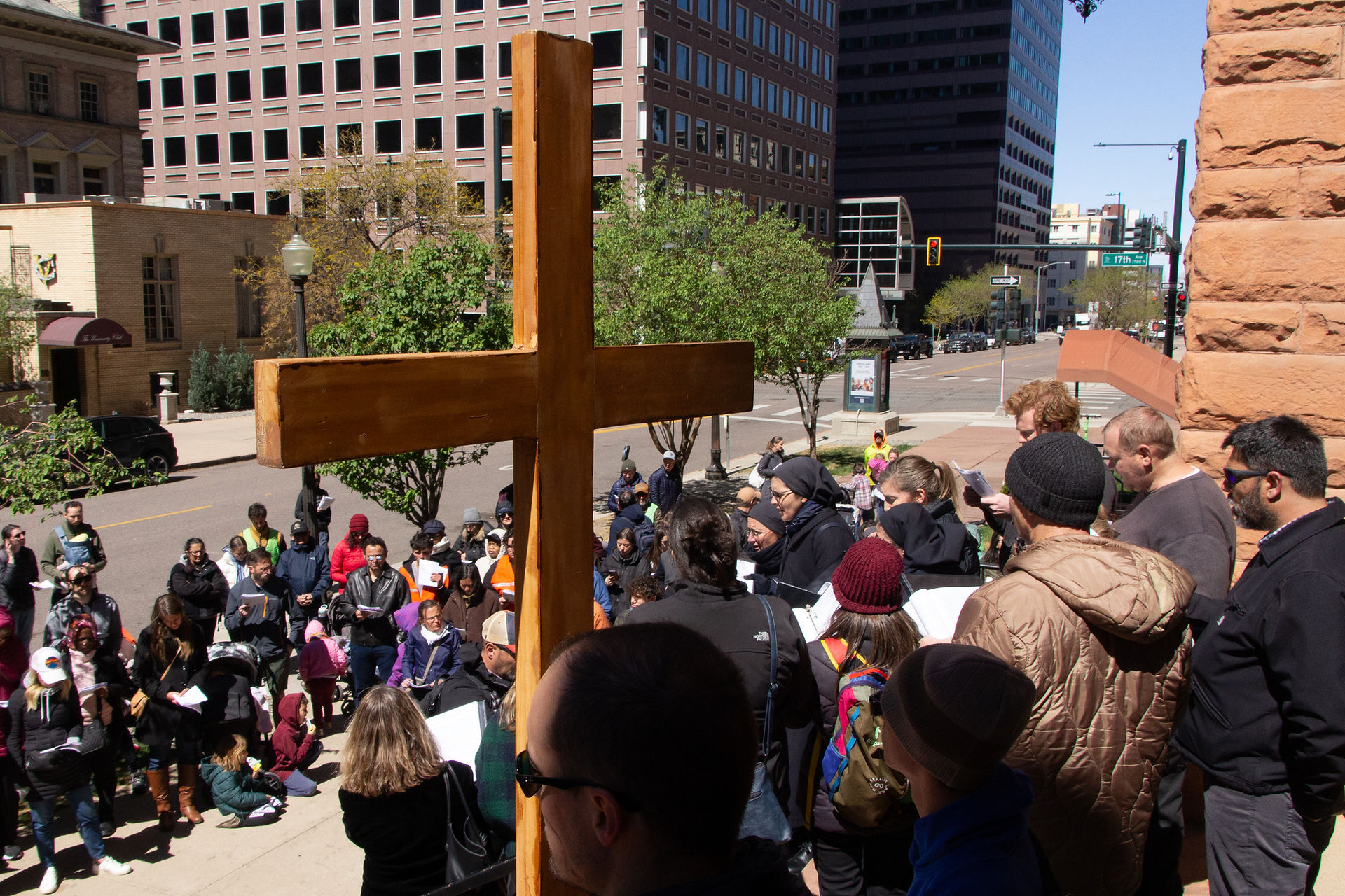 A crowd gathers in a small urban park around a statue while holding prayer sheets during a public devotion.
