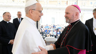 Two clergy members in white and black vestments shake hands, smiling, in an outdoor setting with an audience in the background.