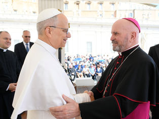 Two clergy members in white and black vestments shake hands, smiling, in an outdoor setting with an audience in the background.