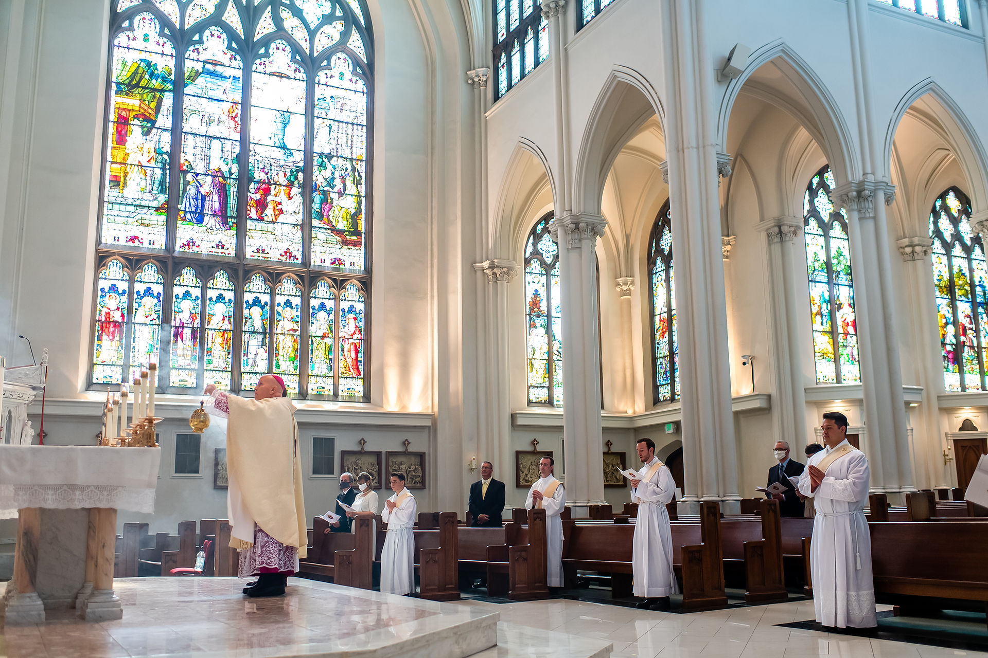 Priest swings incense in ornate cathedral with large stained glass windows. Several people in white robes and suits observe. Sacred atmosphere.