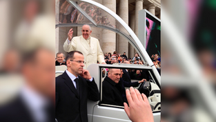 A man in white waves from a vehicle among a crowd. Security personnel stand nearby. Background shows a colonnade and onlookers.