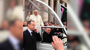A man in white waves from a vehicle among a crowd. Security personnel stand nearby. Background shows a colonnade and onlookers.