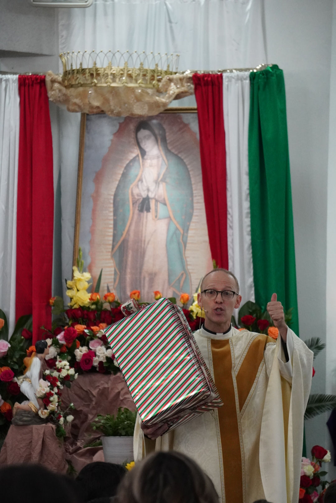 Priest giving a thumbs up, holding a striped gift in a church. Religious art, flowers, and festive drapes are in the background.