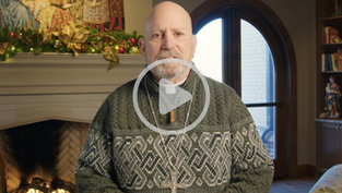Bishop wearing a patterned green sweater and a cross necklace speaks indoors in front of a lit fireplace decorated with Christmas greenery, with bookshelves and an arched window behind him.