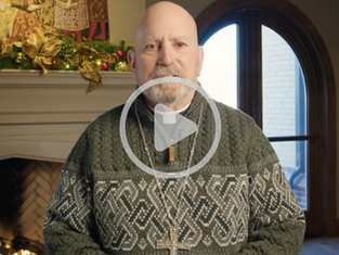 Bishop wearing a patterned green sweater and a cross necklace speaks indoors in front of a lit fireplace decorated with Christmas greenery, with bookshelves and an arched window behind him.