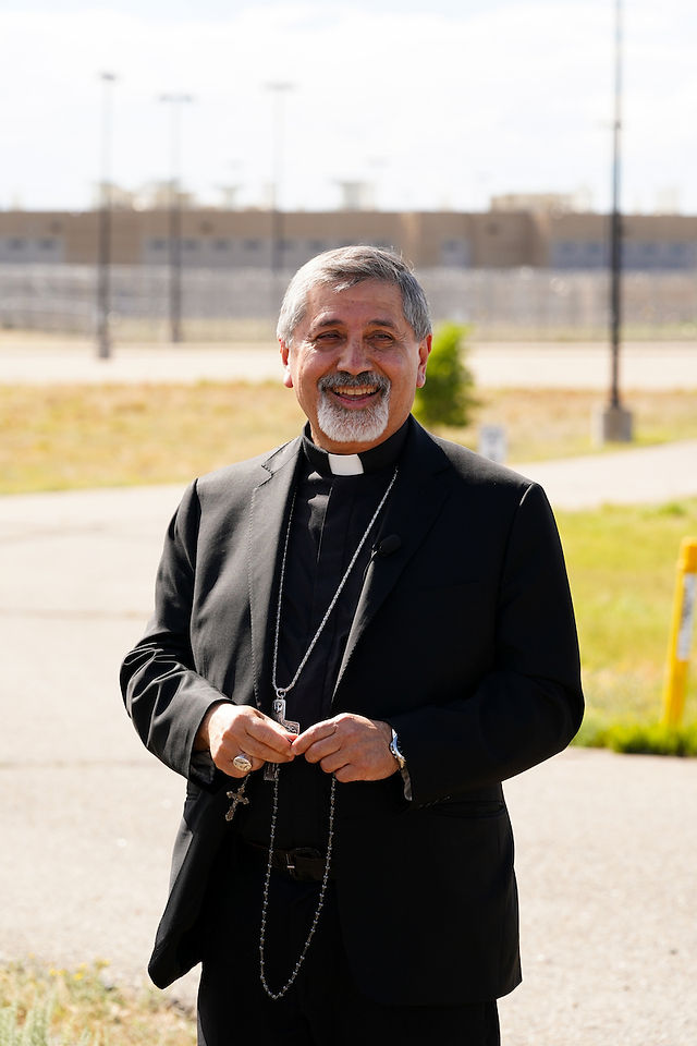 Smiling Catholic priest in black clerical suit and rosary stands outdoors on a sunny path near a fenced building.