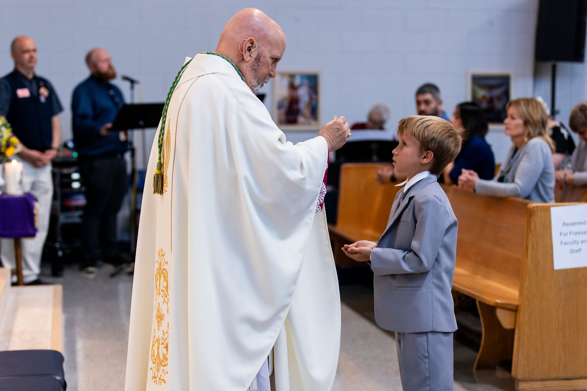 Priest in white robe giving communion to boy in gray suit in a church. Congregation seated, banners visible. Ceremony atmosphere.