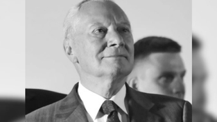 Elderly man in a suit gazes upward with a thoughtful expression. Blurred figure in the background. Black and white image.