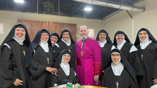 A group of smiling nuns in black habits and a man in a purple robe pose together indoors by a table with soda cans and flowers.