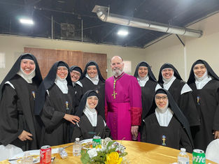 A group of smiling nuns in black habits and a man in a purple robe pose together indoors by a table with soda cans and flowers.
