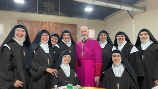 A group of smiling nuns in black habits and a man in a purple robe pose together indoors by a table with soda cans and flowers.