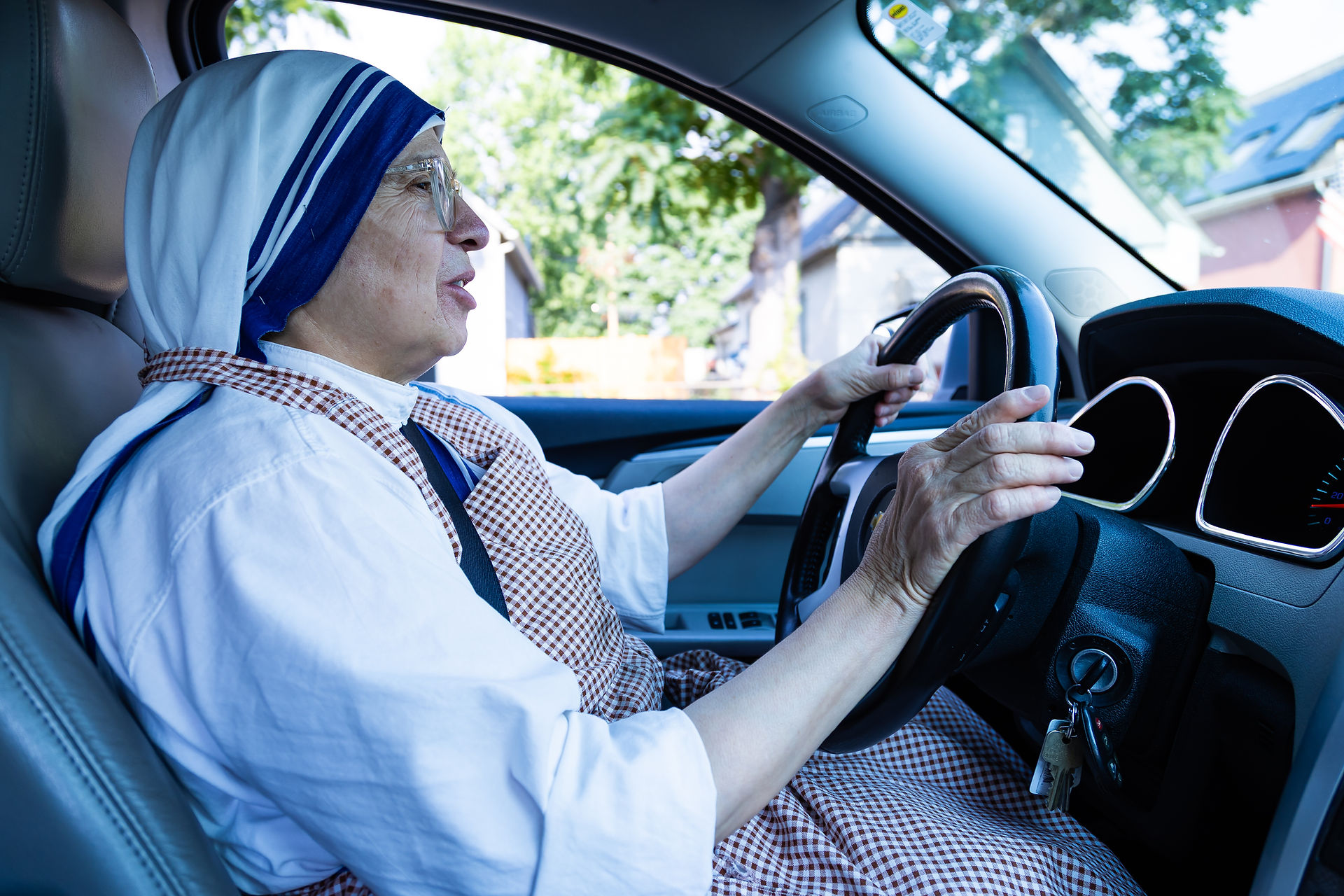 A woman drives a car, wearing a white and blue habit and a checkered apron. Sunlight filters through trees outside the window.