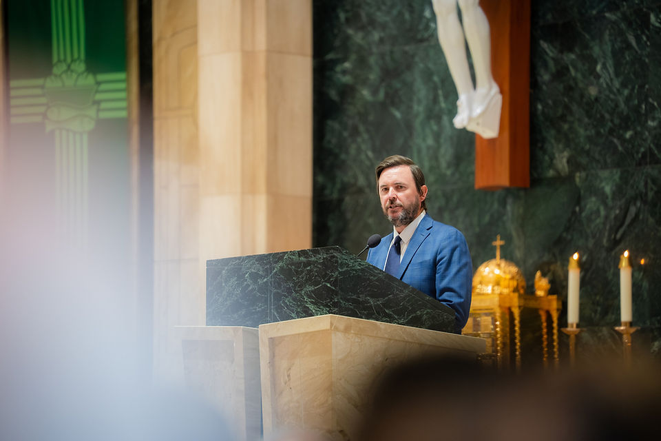 Man in a blue suit speaks at a marble lectern in a church, with candles and a crucifix behind him.