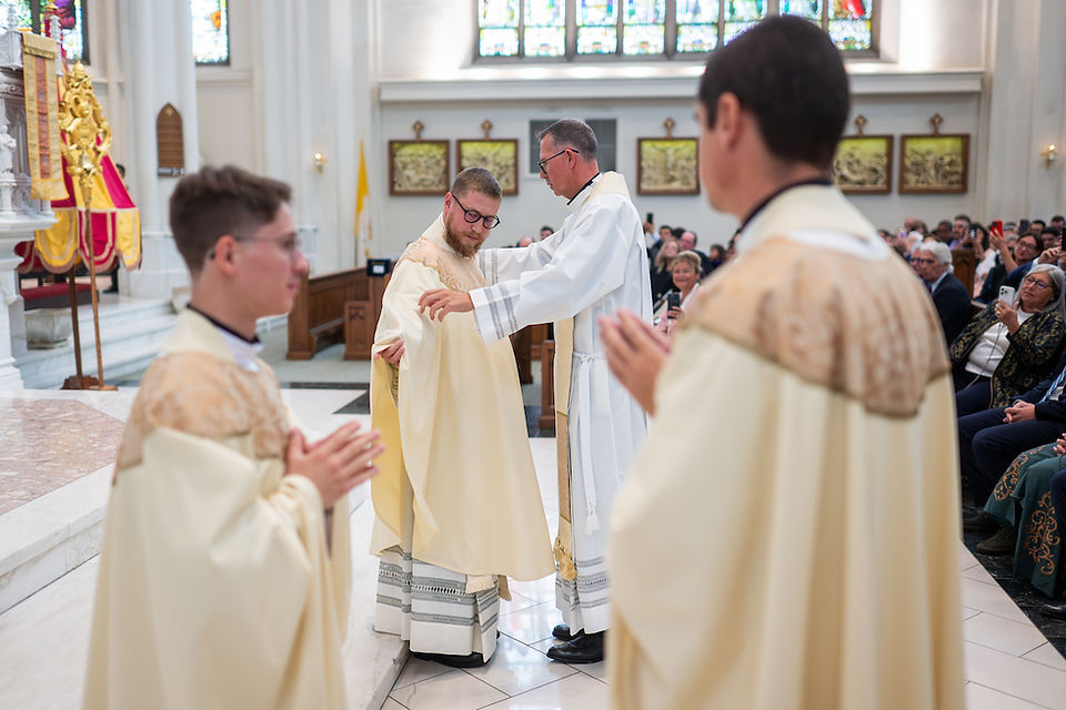 Priests in cream robes perform a ceremony in a church. One adjusts another's robe, while a seated congregation observes. Stained glass above.