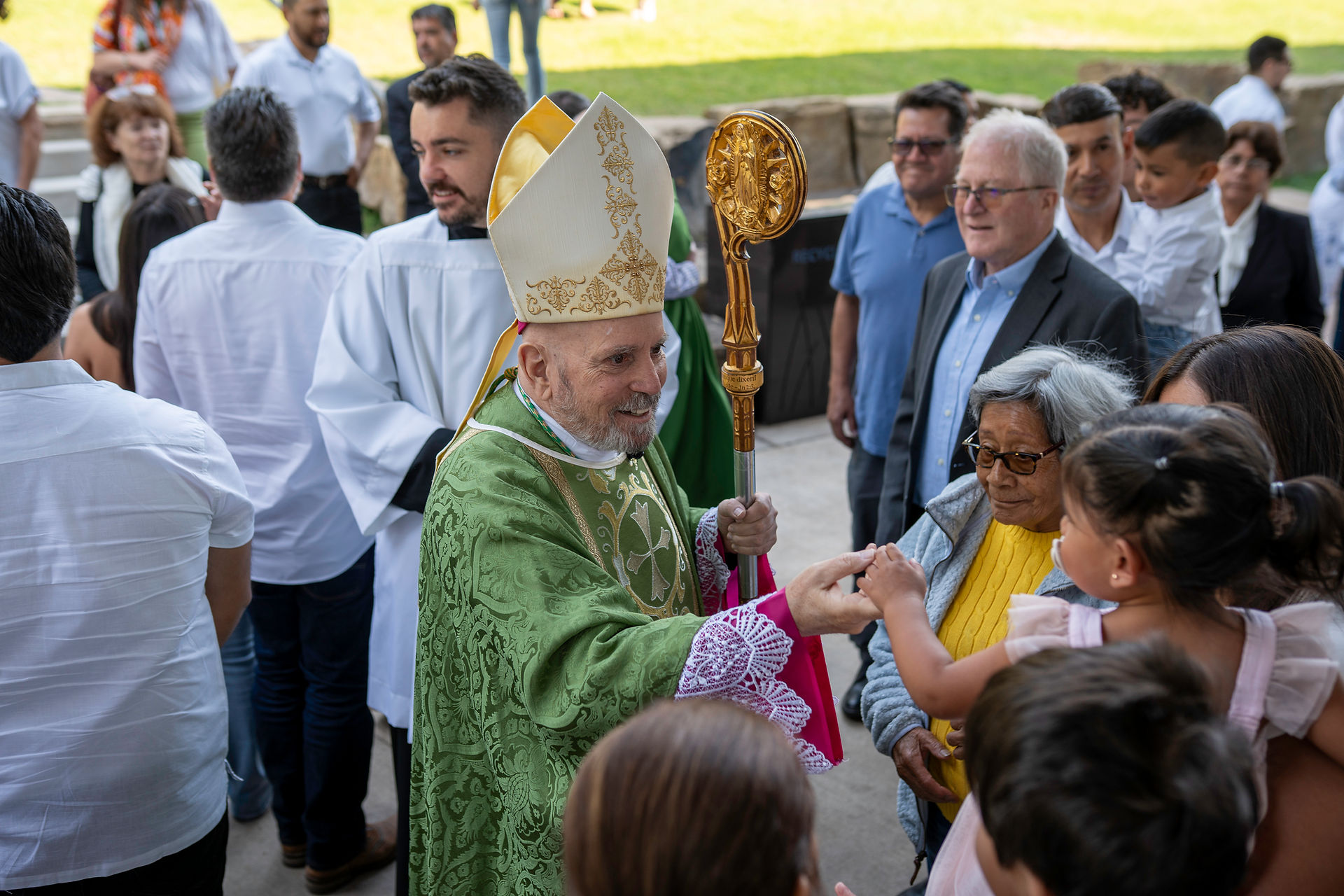 A bishop in green robes and a mitre greets people outdoors. He smiles warmly while holding a ceremonial staff. A crowd surrounds him.