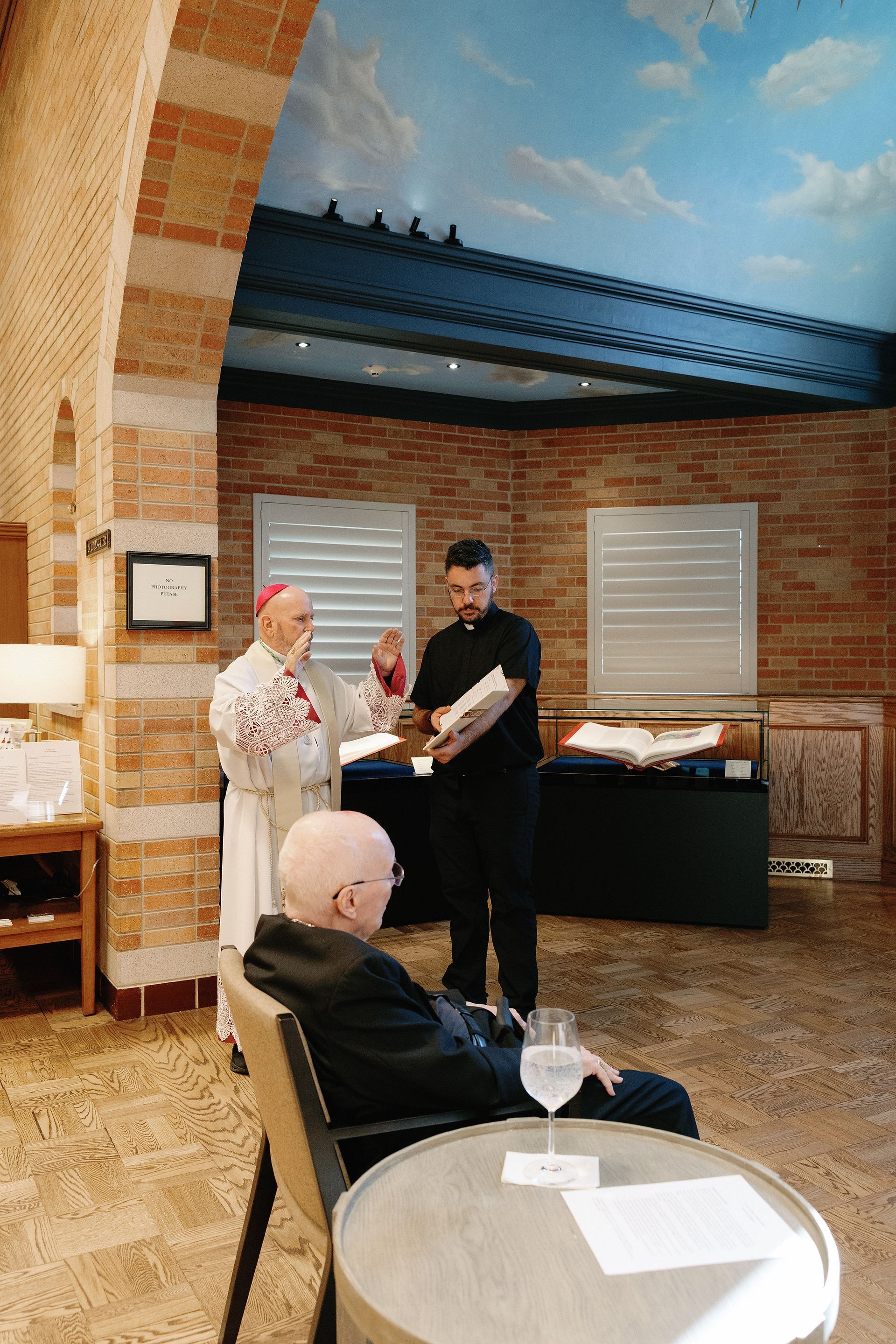 Cleric gestures in discussion with a man holding a book. Elderly man sits at a round table in a warmly lit room with a painted sky ceiling.