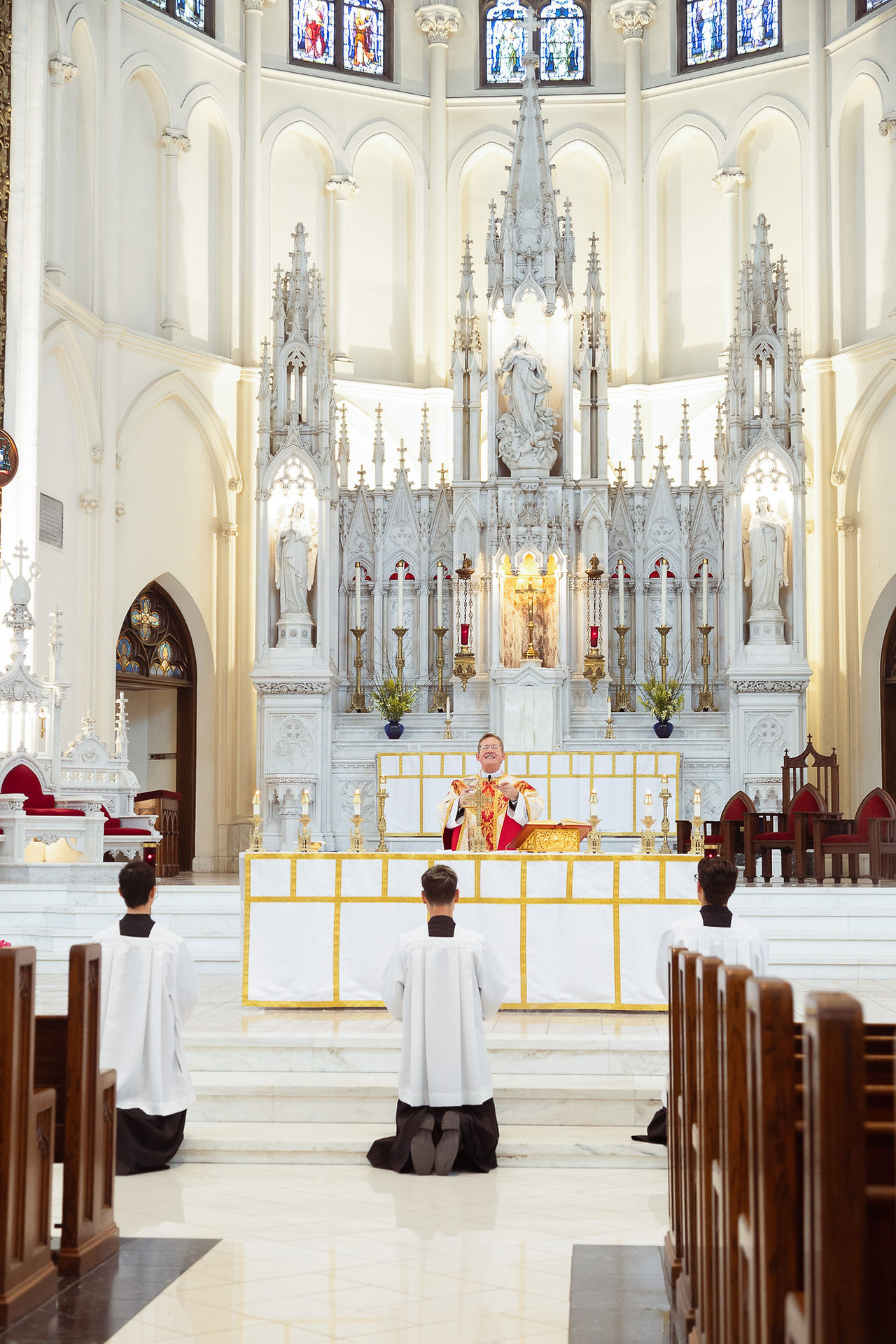 Priest in red vestments leads a service at an ornate altar in a cathedral. Three altar servers kneel in white robes. Stained glass above.