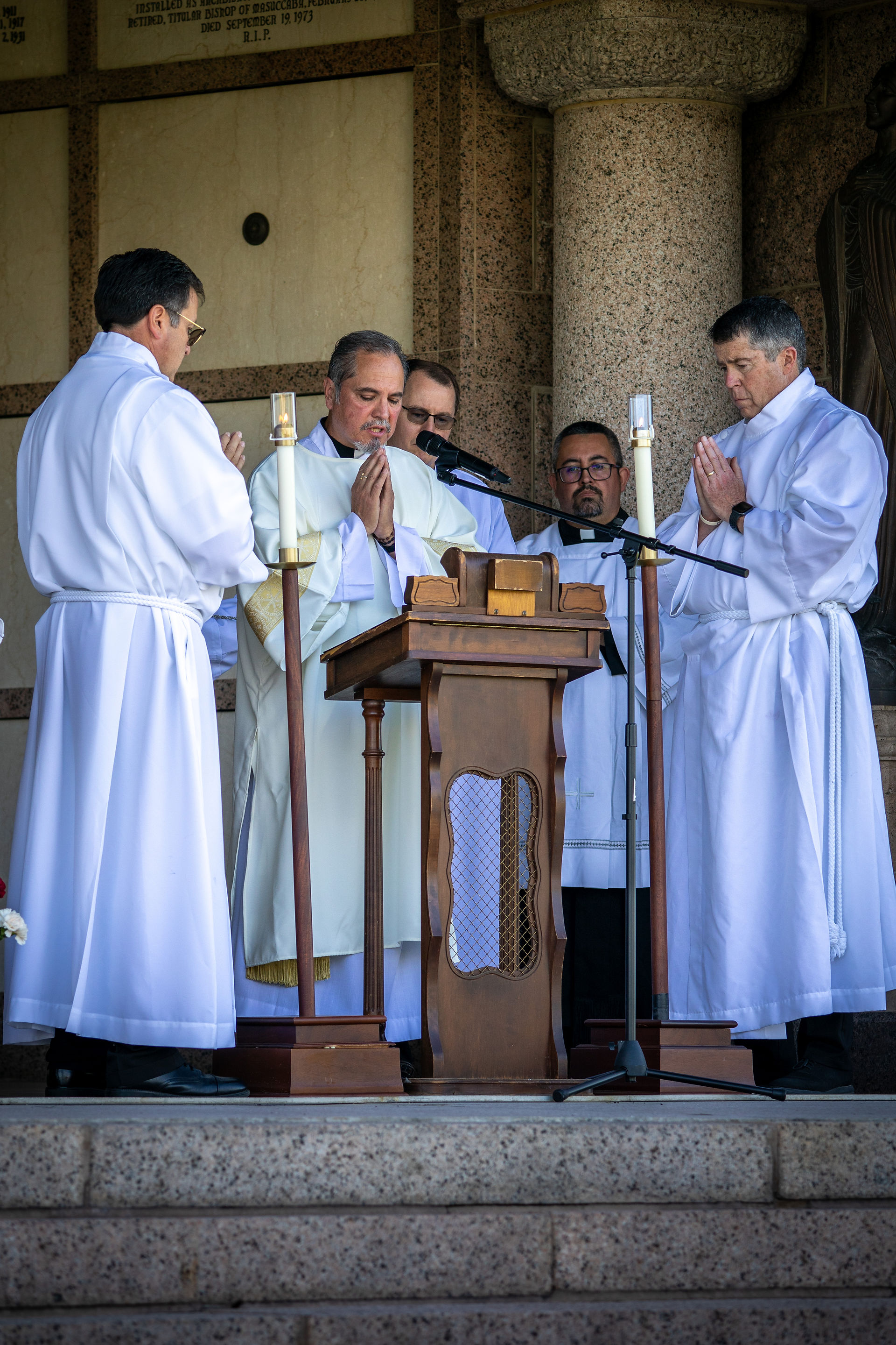 Five men in white robes gather solemnly around a wooden lectern with microphones, in a stone setting, during a ceremony.