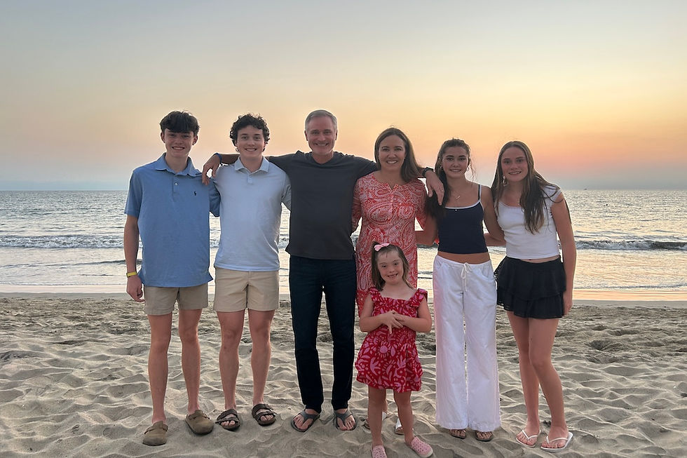 A family of six stands on a sandy beach at sunset, smiling. The ocean waves are visible in the background, creating a warm, serene scene.