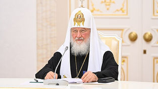 Religious figure in ornate robes sits at a white table with a microphone, holding documents. Ornate interior with gold decor in the background.