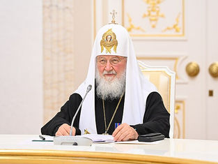 Religious figure in ornate robes sits at a white table with a microphone, holding documents. Ornate interior with gold decor in the background.