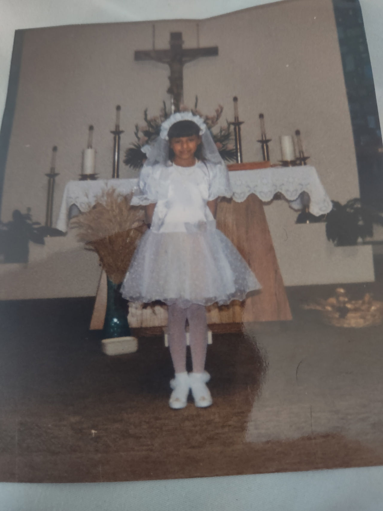 Young girl in a white dress and veil stands smiling in a church, in front of an altar with candles and a crucifix.
