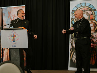 Two clergy members laughing on stage, one at a podium with a cross emblem. Background shows religious artwork, creating a joyful mood.