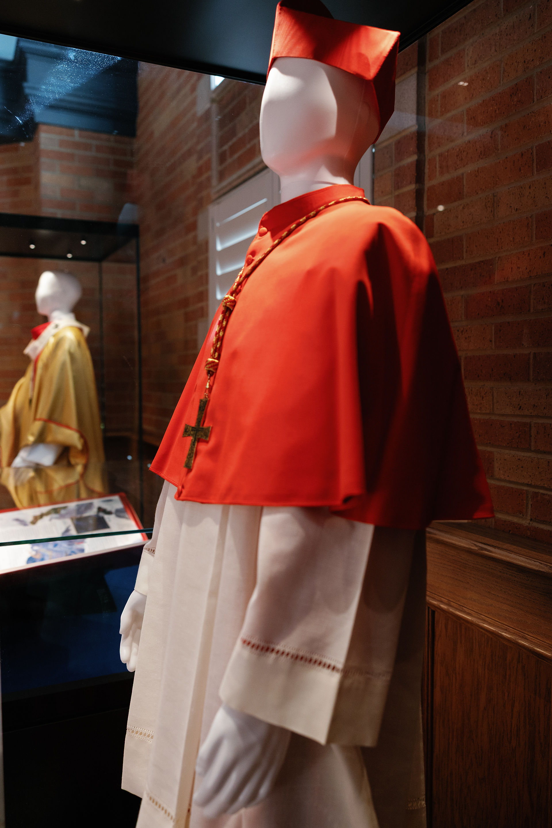 Mannequins in religious attire; one in red with cross necklace, another in yellow. Displayed in a brick-walled museum setting.