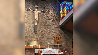 Church altar with a crucifix on a stone wall, stained glass, candles, and a floral cloth. Statues and golden decor add a solemn mood.
