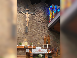 Church altar with a crucifix on a stone wall, stained glass, candles, and a floral cloth. Statues and golden decor add a solemn mood.