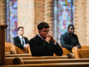 Men in suits sit on pews in a church with stained glass windows, hands clasped in prayer, creating a solemn atmosphere.