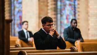 Men in suits sit on pews in a church with stained glass windows, hands clasped in prayer, creating a solemn atmosphere.