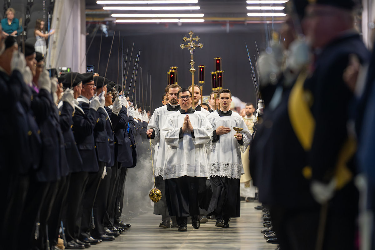 Procession of clergy in white robes holding incense and a cross. Flanked by uniformed guards with swords. Formal, solemn setting.