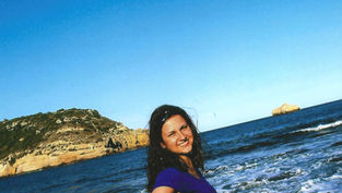 Smiling woman in a blue shirt stands by the ocean, with waves and rocky cliffs in the background under a clear blue sky.