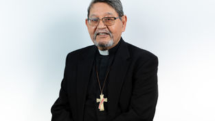 Elderly man in a black suit with clerical collar, wearing glasses and a cross necklace, smiling against a plain white background.