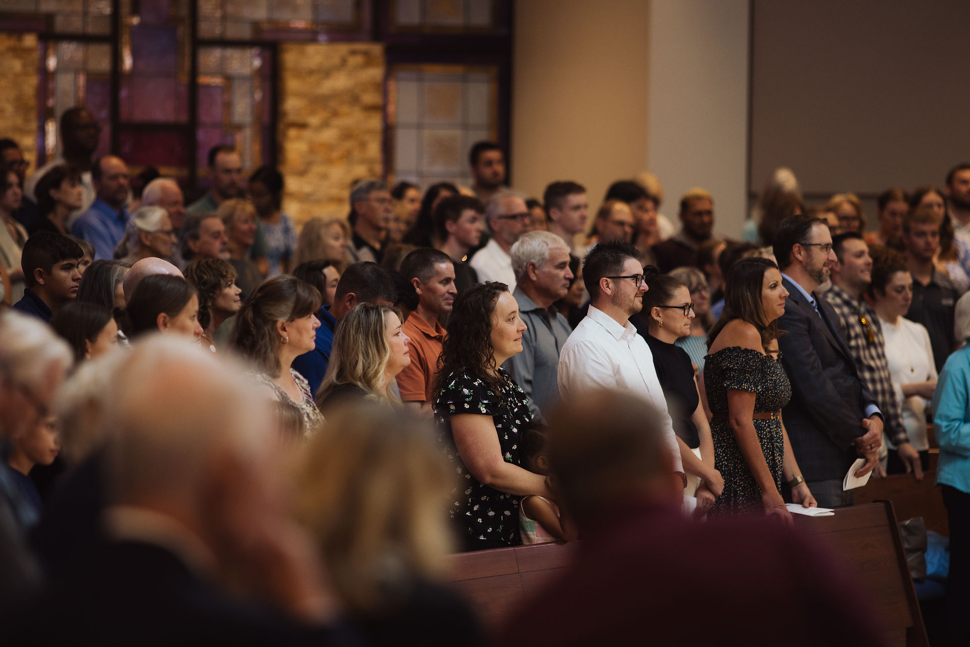 People stand closely in a crowded room, focused and attentive. The backdrop features stained glass and stone, creating a solemn atmosphere.