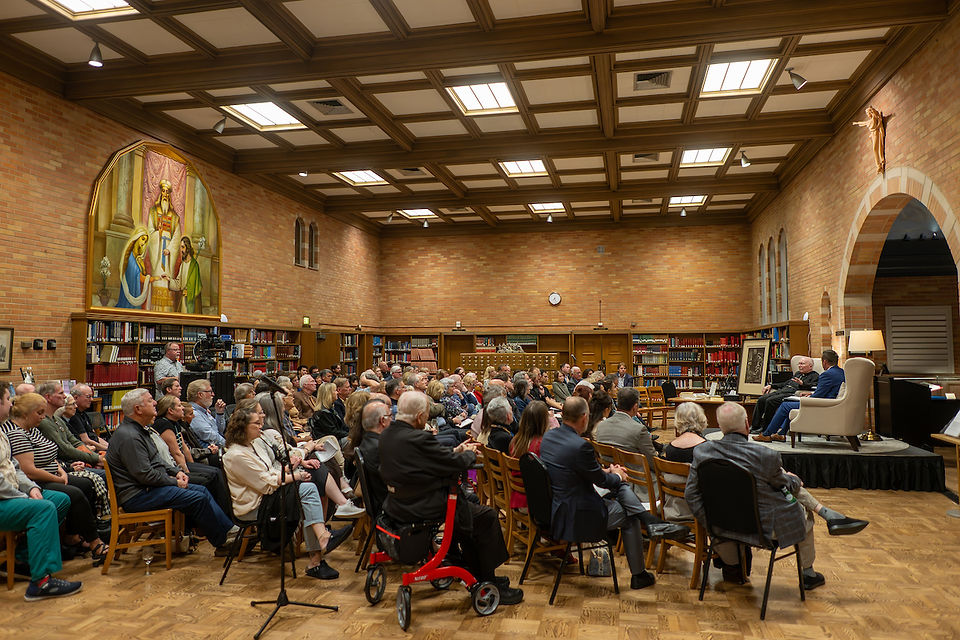 Audience seated in a library with brick walls and a mural, listening to three speakers on stage. Warm lighting creates an inviting atmosphere.