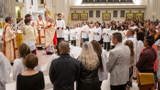 A religious ceremony in a church with a bishop in ornate robes, clergy, and attendees in white robes holding candles, set in a bright interior.