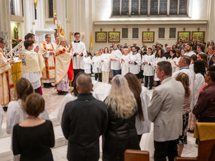A religious ceremony in a church with a bishop in ornate robes, clergy, and attendees in white robes holding candles, set in a bright interior.