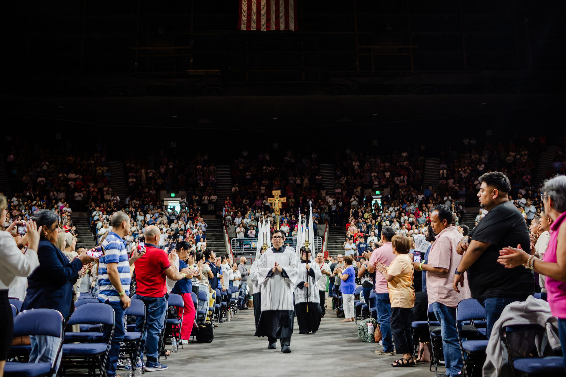 Procession in a crowded arena, led by a person holding a cross. Audience applauds. U.S. flag hangs above. Mood is celebratory.
