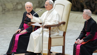 Religious leader in white robe speaks animatedly on a chair, flanked by two clergy in black with pink sashes, in a marble setting.