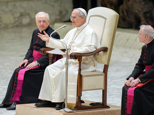 Religious leader in white robe speaks animatedly on a chair, flanked by two clergy in black with pink sashes, in a marble setting.