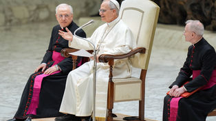 Religious leader in white robe speaks animatedly on a chair, flanked by two clergy in black with pink sashes, in a marble setting.