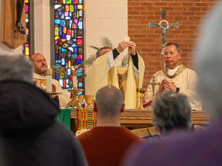 Priest elevates the host during a church service. A colorful stained glass window and attendees in the foreground emphasize reverence.