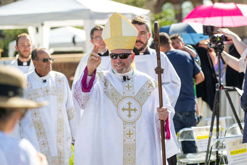 Bishop in white-and-gold vestments and sunglasses blesses a crowd outdoors, holding a staff at a sunny церemony.
