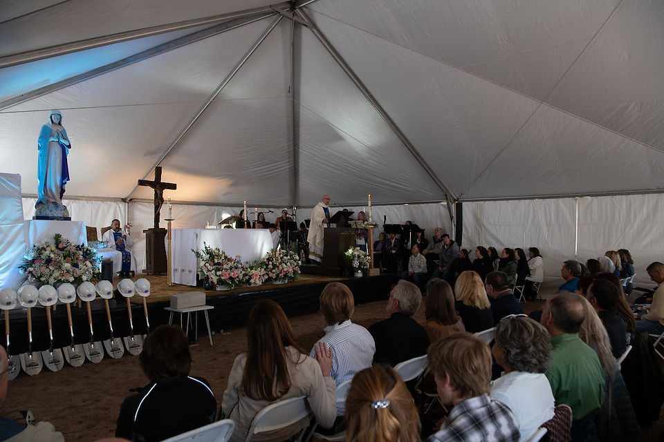 People sit and stand inside a large tent during a liturgical celebration with flowers, candles and a Marian statue near the altar.