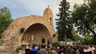 Outdoor gathering at stone grotto with a statue, people seated, speaker at podium, surrounded by trees. Overcast sky, serene atmosphere.