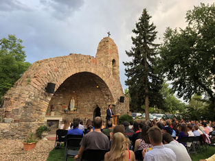 Outdoor gathering at stone grotto with a statue, people seated, speaker at podium, surrounded by trees. Overcast sky, serene atmosphere.